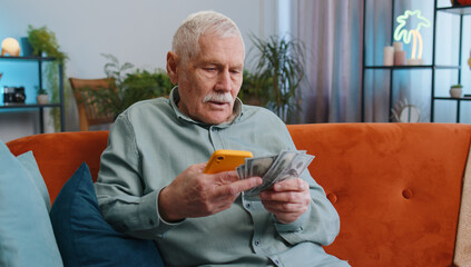 Planning family budget. Senior grandfather man counting money cash, use smartphone calculate domestic bills at home. Joyful elderly guy satisfied of income and saves money for planned vacation, gifts