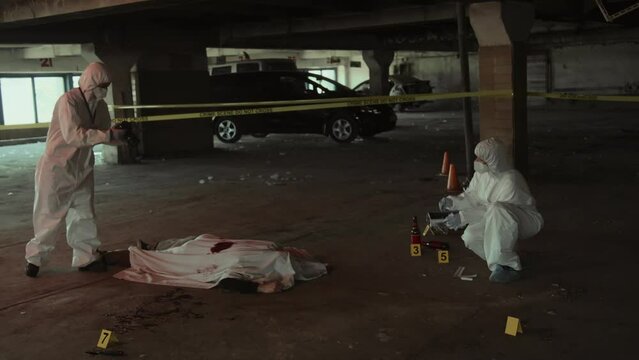 Full shot of two forensic detectives in white protective suits and masks working together at crime scene - talking, taking photographs of corpse covered with bloodied sheet, and searching for clues