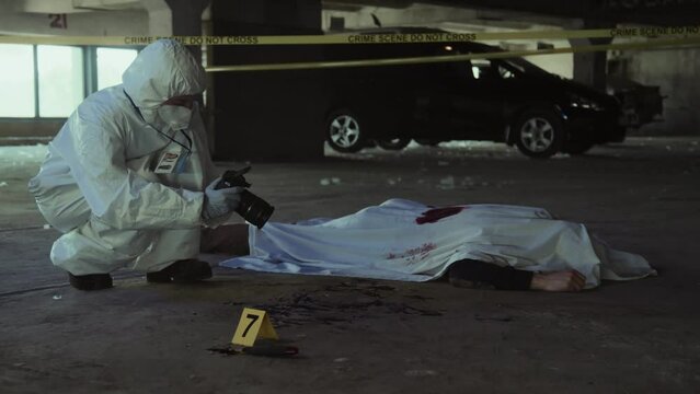 Full shot of unrecognizable male or female forensic investigator in overalls crouching by body of murder victim covered with blood-soaked sheet at disused parking lot, and taking photographs on camera