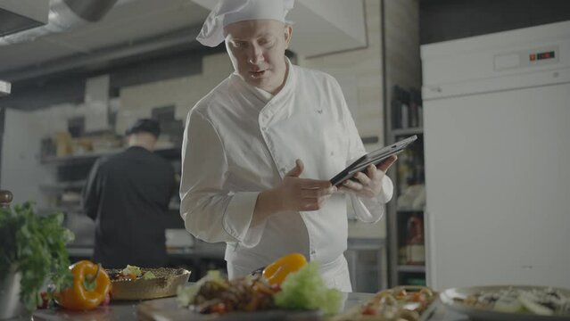 Male chef in white uniform with tablet in his hands checks availability of products on table with list in menu. Slow motion