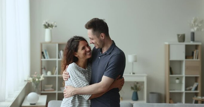 Dreamy calm Hispanic wellbeing spouses hugging standing in living room smile look at camera, daydreaming, cuddling feeling love, express caress enjoy first day at new own apartment. Happy marriage