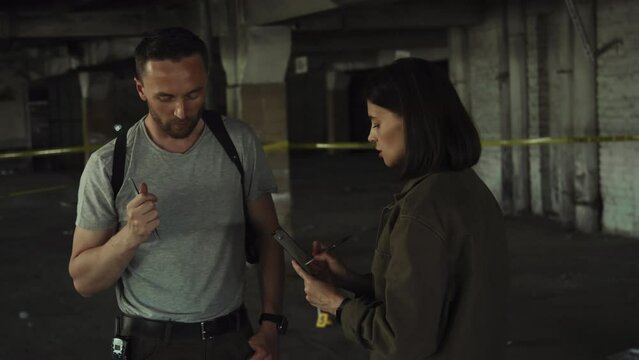 Medium shot of two male and female police operatives talking at crime site - detective in tshirt, gun harness, with portable radio speaking, and colleague with clipboard listening and writing