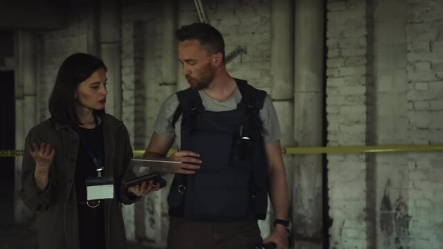 Medium tilting follow shot of Caucasian male police detective in bulletproof vest, with torch walking through murder scene in underground parking, and talking to female colleague with ID badge
