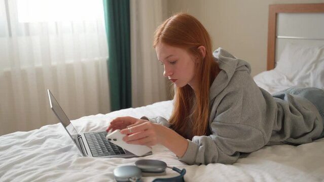 Young woman talking on smartphone while using laptop at home on bed. Teenage girl speaking with friends mobile phone working computer, studying typing, online shopping, watching video, reels