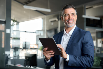 Happy middle aged business man ceo wearing suit standing in office using digital tablet. Smiling...