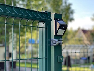 A solar-powered LED street lamp, hanging on a metal fence, in a country cottage, in the summer afternoon. On a blurry background, a village house, trees. The concept of green energy.