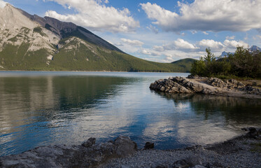 Mountain lake landscape panorama wihout people - 
coniferous forest - pine trees, scenic blue lake, cloudy sky and Rocky Mountains on horizon background. Minnewanka lake, Banff provincial park, Albert