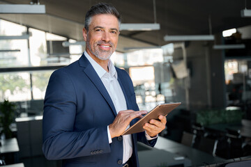 Happy middle aged business man ceo wearing suit standing in office using digital tablet. Confident mature businessman professional company executive looking at camera holding tech device, portrait. - obrazy, fototapety, plakaty
