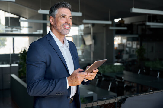Happy middle aged business man ceo wearing suit standing in office using digital tablet. Smiling mature businessman professional executive manager looking away thinking working on fintech device.