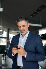 Happy mid aged business man ceo wearing blue suit standing in office using cell phone. Mature businessman professional executive manager holding mobile checking applications on cellphone. Vertical
