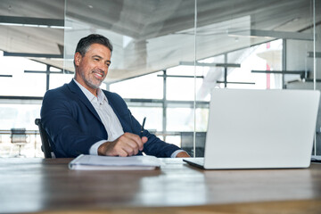 Smiling mature adult business man executive sitting at desk using laptop. Happy professional mid aged businessman ceo manager working on computer technology looking at laptop in office. - obrazy, fototapety, plakaty
