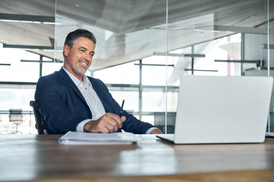 Smiling mature adult business man executive sitting at desk using laptop. Happy professional mid aged businessman ceo manager working on computer technology looking at laptop in office.