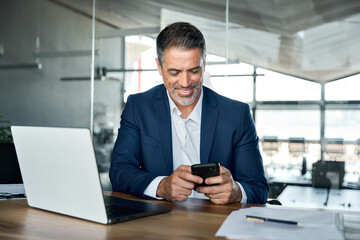 Happy mid aged business man ceo executive, financial expert investor wearing suit sitting in modern office holding smartphone using mobile phone managing digital tech work transactions at workplace.