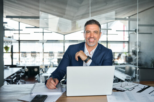 Happy middle aged professional business man company executive ceo manager wearing suit sitting at desk in modern office working on laptop computer and writing notes, portrait at workplace.
