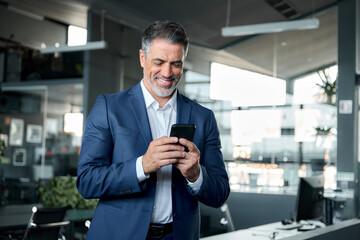 Fototapeta na wymiar Happy middle-aged business man ceo wearing blue suit standing in office using cell phone. Older businessman professional executive holding mobile satisfied with enterprise solution management service.