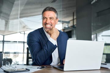 Happy smiling middle aged professional business man company executive ceo manager wearing suit sitting at desk in office working on laptop computer laughing at workplace. Authentic candid photo. - obrazy, fototapety, plakaty