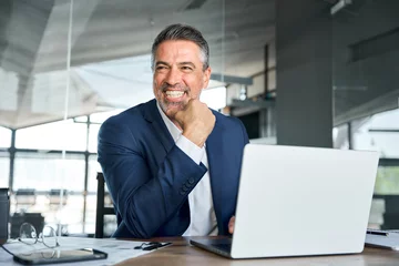 Fotobehang Happy smiling middle aged professional business man company executive ceo manager wearing suit sitting at desk in office working on laptop computer laughing at workplace. Authentic candid photo. © insta_photos