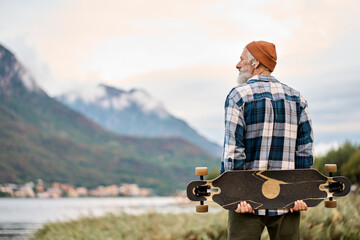 Active cool happy bearded old hipster man standing in nature park holding skateboard. Mature traveler skater enjoying freedom spirit and extreme sports hobby looking at mountains view. - Powered by Adobe