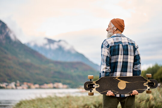 Active cool happy bearded old hipster man standing in nature park holding skateboard. Mature traveler skater enjoying freedom spirit and extreme sports hobby looking at mountains view.