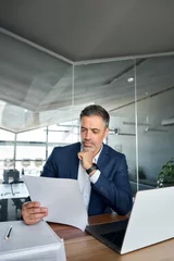 Fotobehang Lengtemeter Busy middle aged professional business man lawyer or financial law expert wearing suit holding corporate documents reading paper contract sitting at desk in office managing deal risks. Vertical