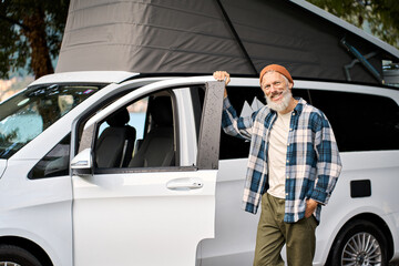 Happy older man standing with rv camper van on travel vacation. Smiling mature active traveler advertising campervan car with tent on roof enjoying camping tourism road trip, campervan booking rental.