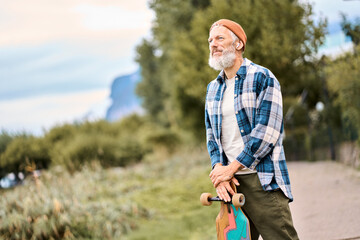 Active cool bearded old hipster man standing in nature park holding skateboard. Mature traveler skater enjoying freedom spirit and extreme sports hobby leisure lifestyle, authentic shot.