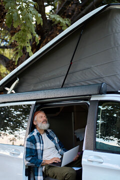 Calm Older Man Sitting In Rv Camper Van Using Laptop. Peaceful Mature Traveler Holding Computer On Lap Remote Working Online And Enjoying Vanlife, Freedom, Resting In Outdoor Camping. Vertical