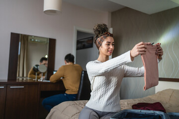 Woman caucasian couple packing or unpack wardrobe cloth from suitcase at hotel room while her husband or boyfriend sit in background happy smile travel and vacation concept copy space