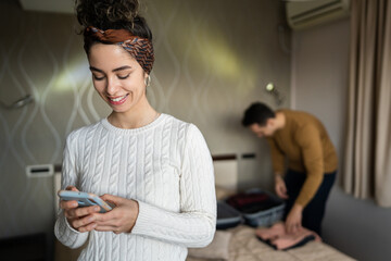 couple young beautiful woman and man in hotel room wife or girlfriend use mobile phone for texting sens sms messages while husband or boyfriend is unpacking or packing baggage on the bed copy space - obrazy, fototapety, plakaty