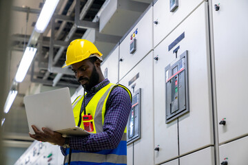 Professional African male engineer in safety uniform working at factory server electric control panel room. Industrial technician worker maintenance checking power system at manufacturing plant room.