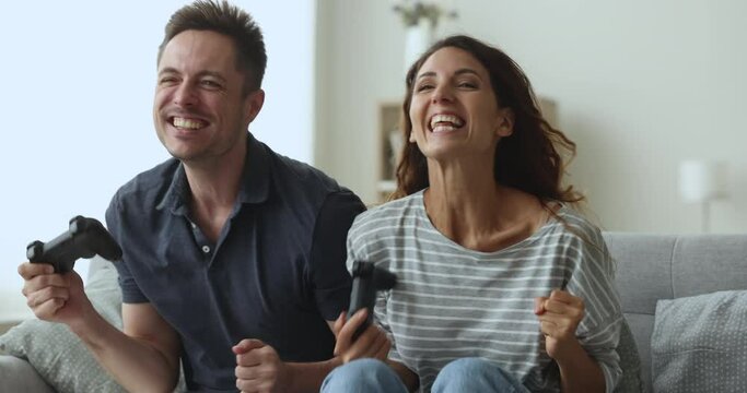 Smiling wife and husband spend family weekend at home sit on couch in living room, play video game on electronic station combat in team at virtual world, feel happy giving high five, celebrate victory