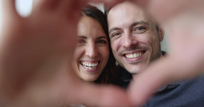 Close up overjoyed, affectionate Hispanic couple looking at camera through joined fingers showing heart symbol. Romantic spouses shoot video, make selfie, celebrate St. Valentine Day. Love, relations