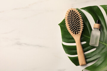 Wooden hair brush, bottle of essential oil and monstera leaf on white marble table, top view. Space for text