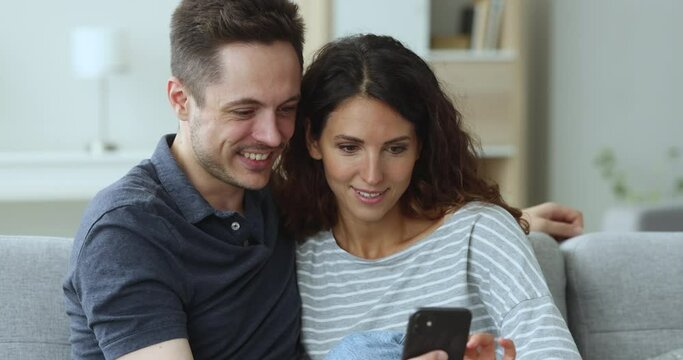 Pretty Hispanic couple use cellphone seated on sofa, look at screen, learn new mobile application, choose goods through e-commerce website, watch videos, review photos spend pastime at home, close up