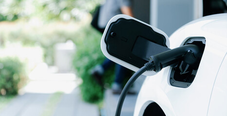 Focus electric car charging at home charging station with blurred progressive man walking in the...