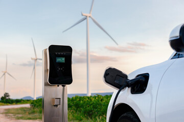 Progressive combination of wind turbine and EV car, future energy infrastructure. Electric vehicle being charged at charging station powered by renewable energy from wind turbine in the countryside. - obrazy, fototapety, plakaty