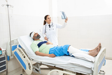 Indian man patient lying on bed with fractured leg and orthopedic doctor looking at foot X-ray film...