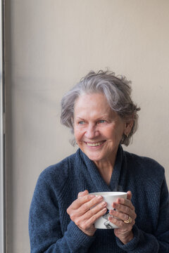 Vertical closeup of senior woman in blue sweater holding tea cup and laughing (selective focus)