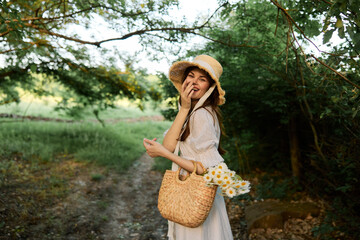 happy, smiling woman in a wicker hat with a basket of daisies in her hands walks through the forest
