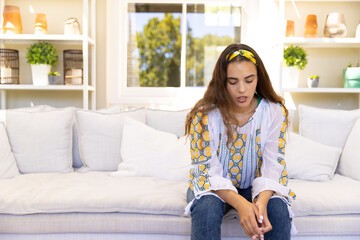 Biracial depressed young woman with long hair sitting on sofa in living room, copy space