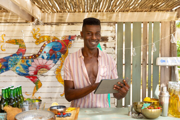 Portrait of african american male bartender holding tablet in beach bar, unaltered