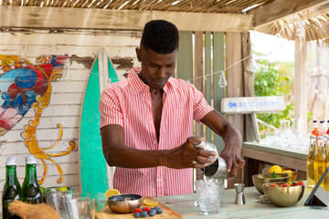 African american male bartender holding shaker and preparing cocktails in beach bar, unaltered