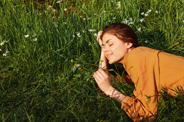a calm woman with long red hair lies in a green field with yellow flowers, in an orange dress, smiling pleasantly, closing her eyes from the bright summer sun, resting her head on her hands