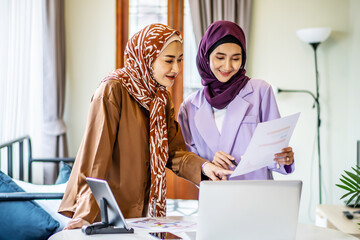 Two Asian muslim women wearing hijab working together with using laptop pc tablet and paper showing graphic chart in living room at home, respect and collaboration concept. - obrazy, fototapety, plakaty