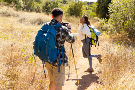 Happy biracial couple wearing backpacks, hiking and taking pictures with camera in forest