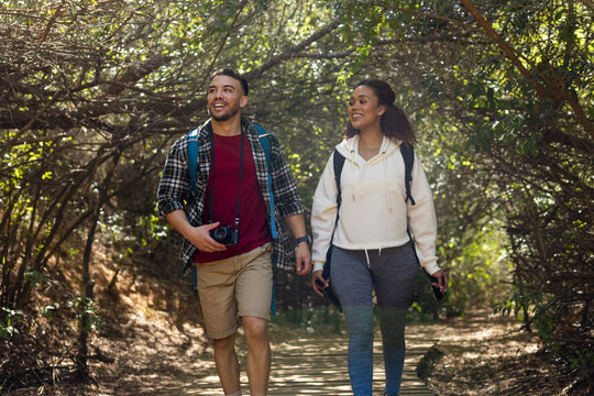 Happy biracial couple wearing backpacks and hiking in forest