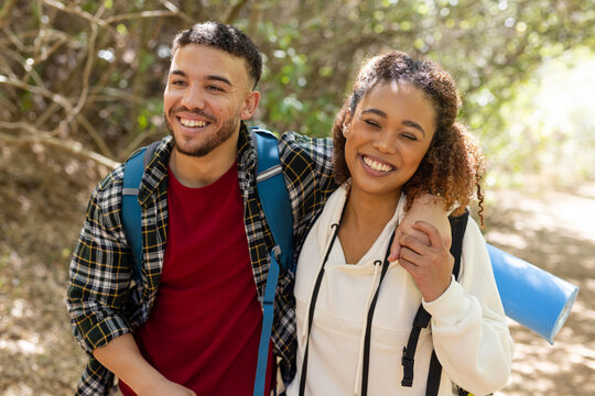 Happy biracial couple wearing backpacks, embracing and smiling in forest