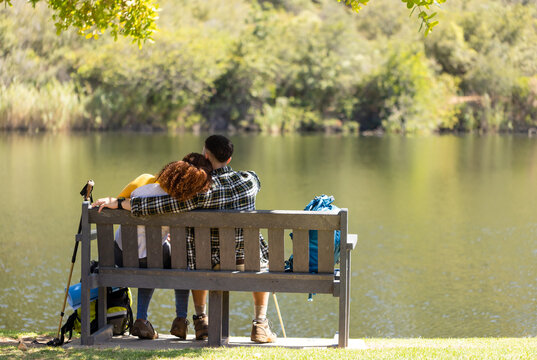 Happy biracial couple sitting on bench and embracing by lake