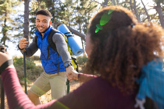 Happy biracial couple wearing backpacks, hiking and holding hands in forest
