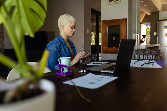 Unaltered biracial casual businesswoman using smartphone in home office with laptop and paperwork